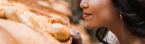 Preview: panoramic shot of cheerful woman smiling while smelling bread in supermarket