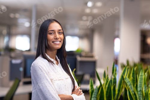 Preview: Portrait of smiling young biracial businesswoman standing with arms crossed at modern workplace