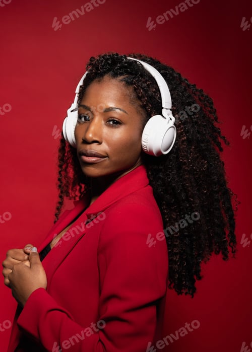Preview: Hispanic woman with curly hair in red blazer wearing headphones listening music on red background