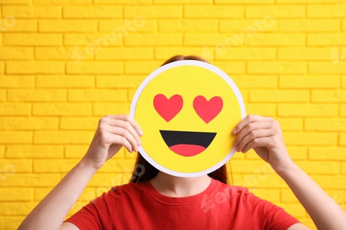 Preview: Woman covering face with heart eyes emoji near yellow brick wall