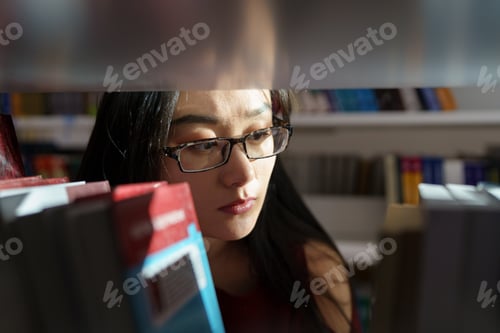 Preview: Asian girl student looking for book or textbook on library bookshelf studying at university campus