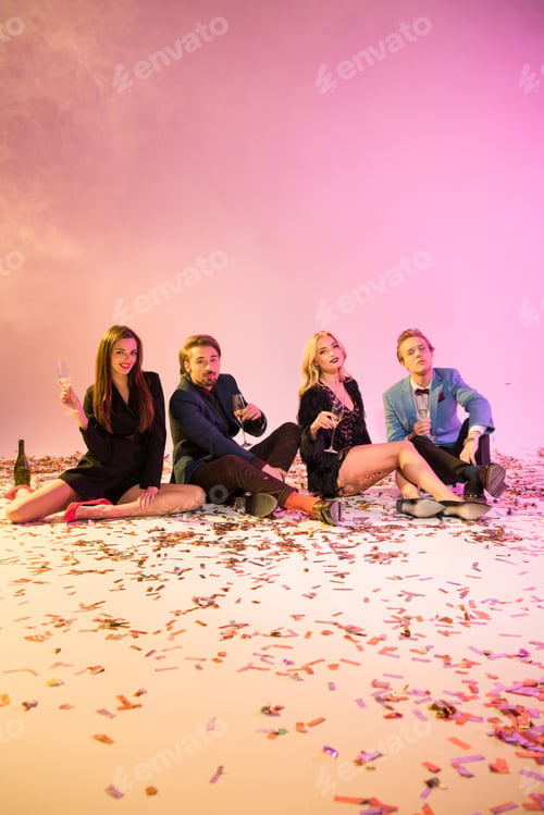 Preview: happy fashionable friends with champagne glasses sitting on floor with confetti, on pink