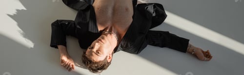 Preview: top view of shirtless man in black blazer and silver necklace lying and looking at camera on grey