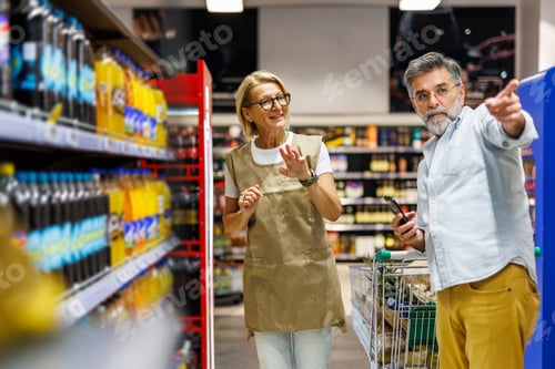 Preview: Senior customer pointing at shelf while shopping with supermarket employee