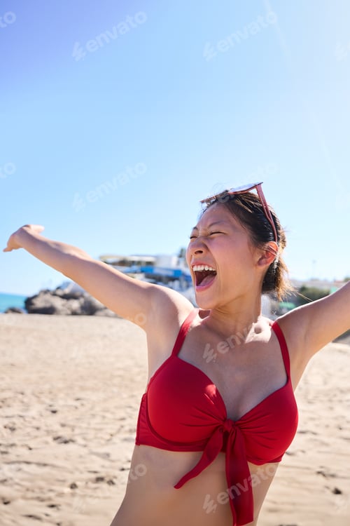 Preview: Portrait of young Asian Chinese woman in red bikini with big smile while raising arms on the beach