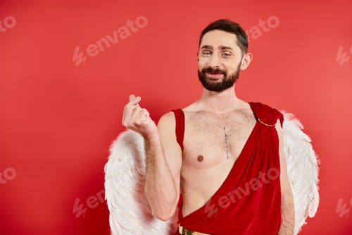 Preview: joyful bearded cupid man showing small heart sign with fingers and looking at camera on red