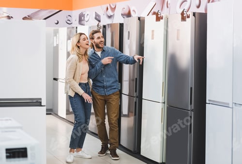 Preview: smiling girlfriend and boyfriend pointing with fingers at fridge in home appliance store