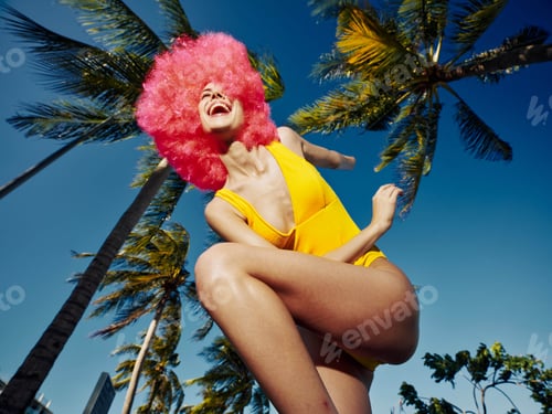 Preview: Yellow swimsuit woman with pink wig jumping in air, tropical palm trees background