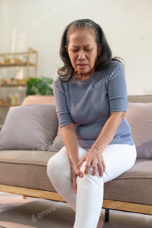 Preview: Senior asian woman experiencing knee pain sitting on sofa at home.