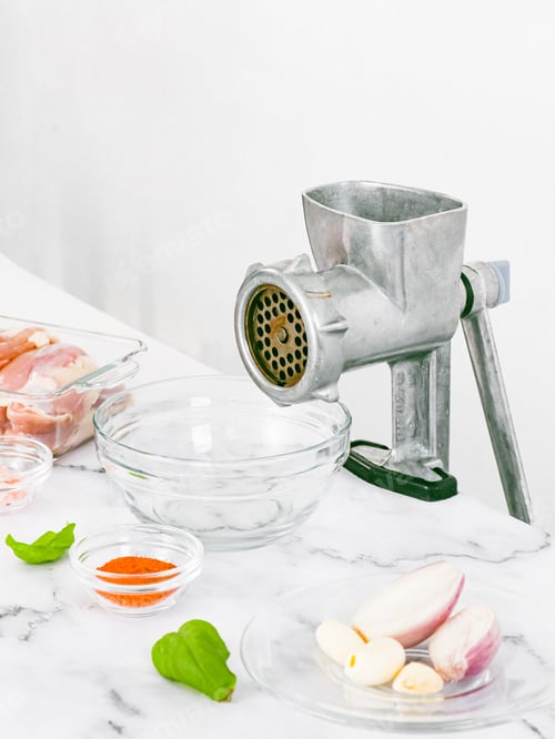 Preview: Manual meat grinder with glass bowl,boneless chicken thighs,peeled onions,garlic on a marble table .
