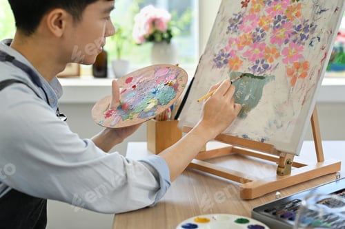 Preview: Image of pleased man artist painting picture with watercolor on canvas in art workshop.