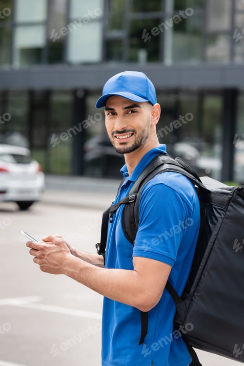 Preview: Young arabian courier with thermo backpack and smartphone looking at camera on urban street