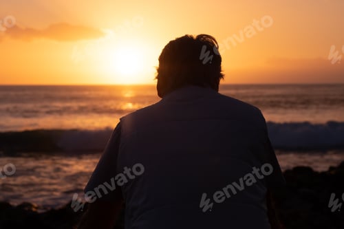 Preview: Rear view of mature man at sea looking at the horizon and at the orange sunset light