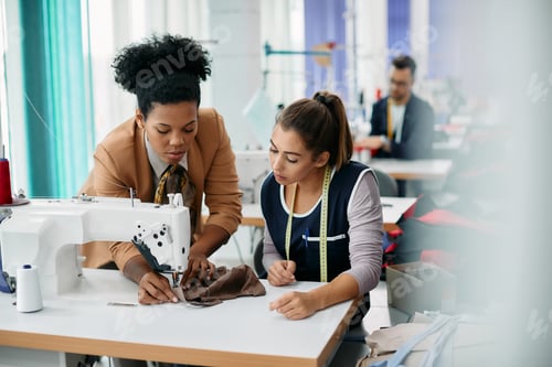 Preview: Black clothing designer assisting young seamstress with sewing in a textile workshop.