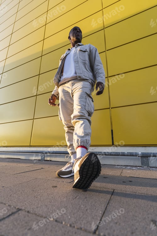 Preview: Vertical low angle shot of a young Black male standing in front of a yellow wall
