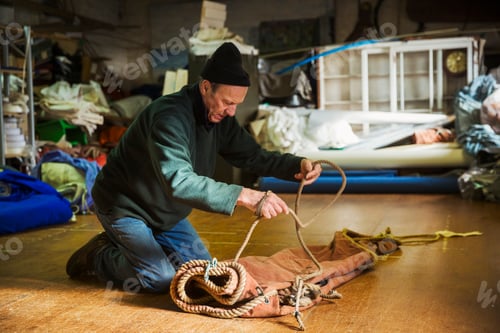 Preview: Man in a sailmaker's workshop kneeling on the floor, tying a rope on a sail.