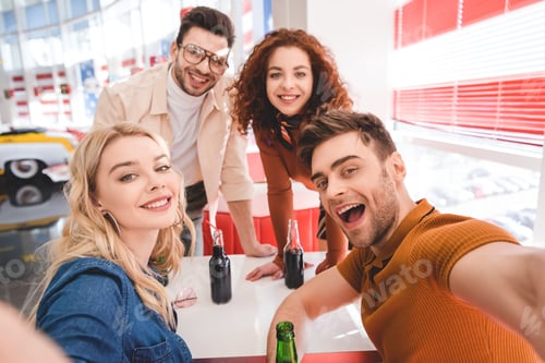 Preview: selective focus of smiling handsome and attractive friends holding glass bottles with soda and beer