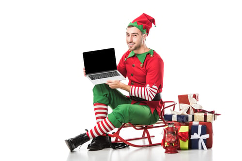 Preview: man in christmas elf costume sitting on sleigh and holding laptop with blank screen isolated on