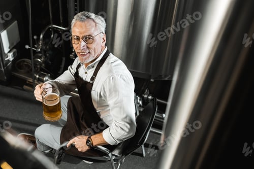 Preview: overhead view of smiling senior brewer in apron holding glass of beer in brewery