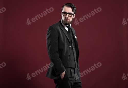 Preview: Retro 1900 Fashion Man With Beard Wearing Grey Suit Black Tie And Glasses. Studio Shot Against Red