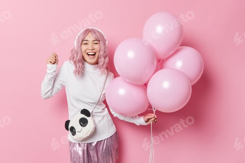 Preview: Positive carefree Asian girl with rosy hair listens music via headphones holds bunch of helium ballo