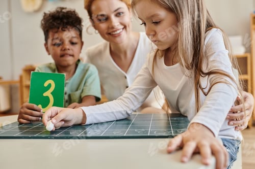 Preview: girl writing on chalkboard near african american boy with number and teacher, Montessori school