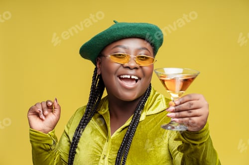 Preview: Happy stylish African American woman drinking alcohol cocktail isolated on yellow background in bar