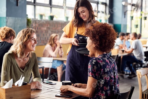 Preview: Two Female Friends Sitting At Table In Coffee Shop Being Served By Waitress