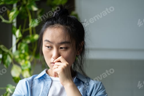 Preview: Worried pensive Asian woman touching lips nervous stands in apartment near closet with houseplants