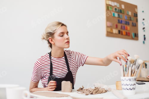 Preview: Girl in black apron and striped T-shirt sitting at the table with clay thoughtfully selecting tools