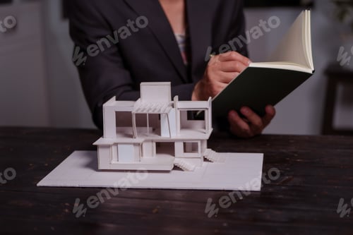 Preview: A businessman sits at a table at night, holding a model of a two-story white house. He reviews