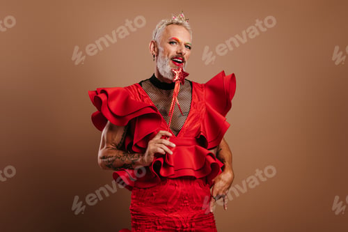 Preview: Playful gay man in beautiful red dress holding magic wand against brown background