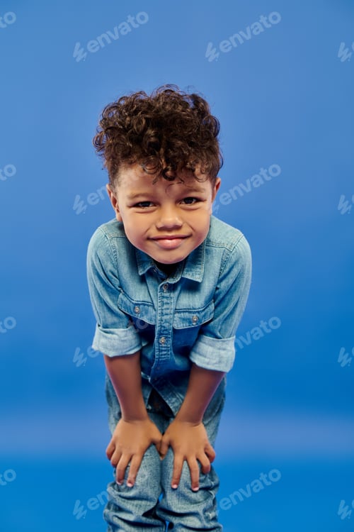 Preview: little happy african american preschooler boy in stylish denim outfit posing on blue background