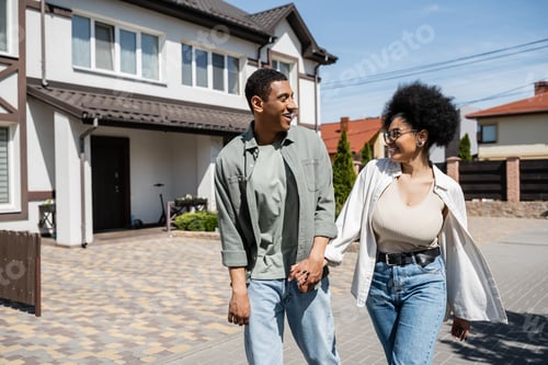Preview: joyful african american couple holding hands while walking on street near houses in summer