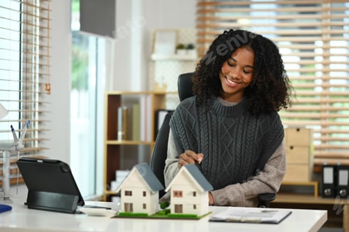 Preview: Smiling African American woman with house model and wind turbine, sustainable energy.