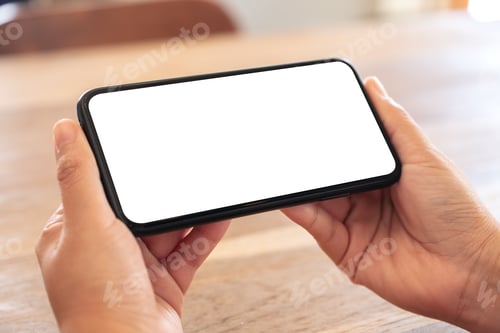 Preview: woman's hands holding black mobile phone with blank white screen horizontally on wooden table