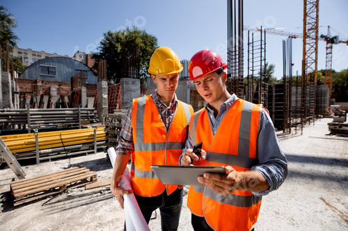Preview: Architect and structural engineer dressed in orange work vests and helmets discuss a building