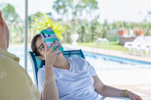 Preview: young latina woman leaning on a chair looking at her cell phone while lazing around on a hot day