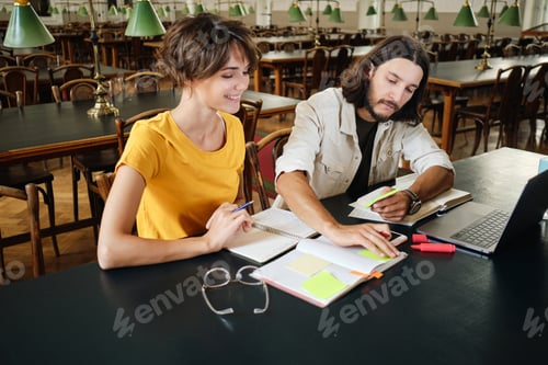 Preview: Two young cheerful students happily making notes studying together in library of university