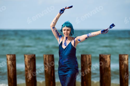 Preview: Artistic blue-haired woman performance artist smeared with blue gouache paints dancing on beach