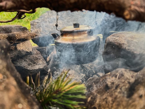 Preview: Blackened kettle heating over campfire. Smoke swirls around kettle, and hint of green pine needles.