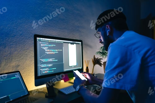 Preview: Young man mobile developer writes program code on a computer and check app in smartphone