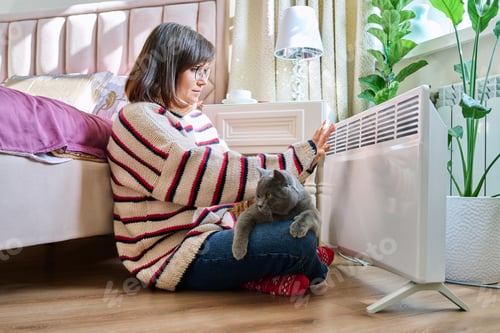 Preview: Woman sitting with cat in a warm sweater warming near an electric heater