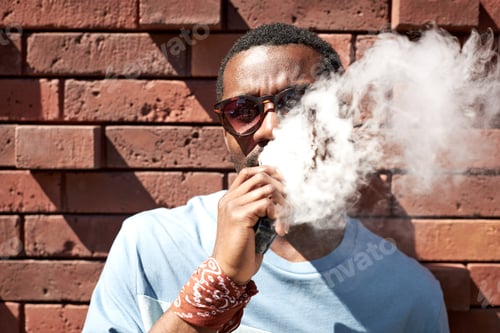 Preview: African American guy in sunglasses and blue t-shirt smoking by brick wall