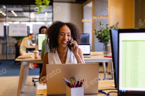 Preview: Happy biracial casual businesswoman using laptop and talking on smartphone in office