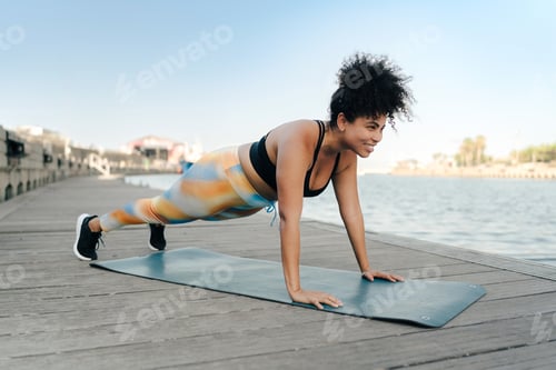 Preview: Curvy hispanic woman doing push-ups on a fitness mat on a wooden pier by the sea
