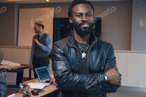 Vista previa: Retrato de un joven africano con rastas y barba con los brazos cruzados dentro del espacio de trabajo conjunto.