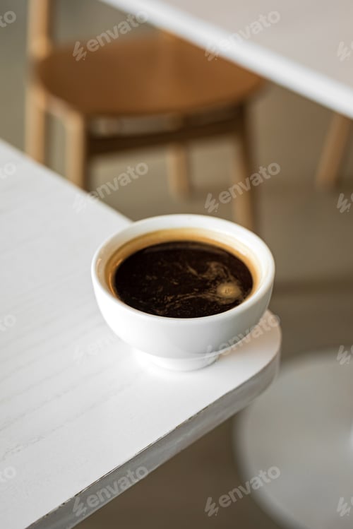 Preview: A white cup of black coffee americano on the edge of a table in a cafe
