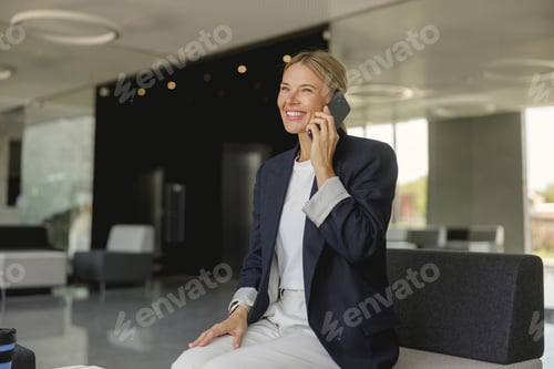 Preview: Cheerful businesswoman talking phone sitting on modern office background and looks away