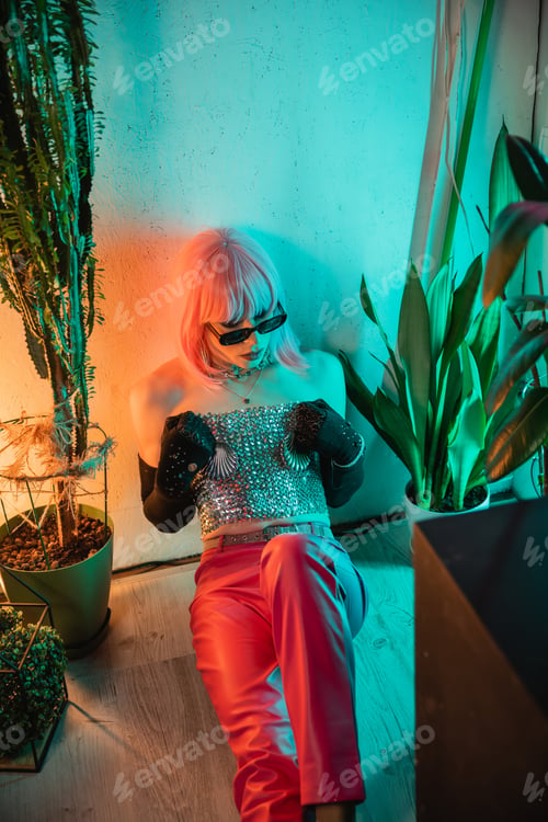 Preview: Fashionable drag queen in sunglasses and silver top sitting near plants at home